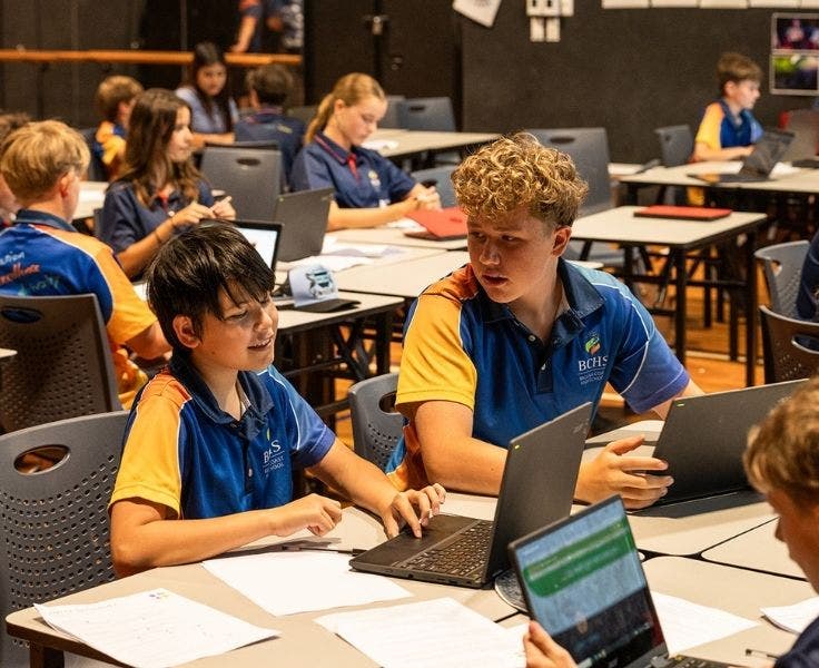 Two young male sudents working together on laptops with other students doing the same thing in the background