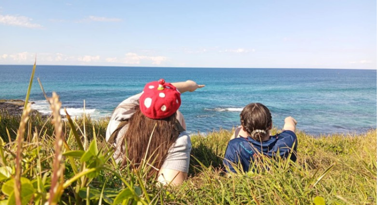 Back view of teacher and student relaxing back on a grassy headland both pointing out towards the ocean