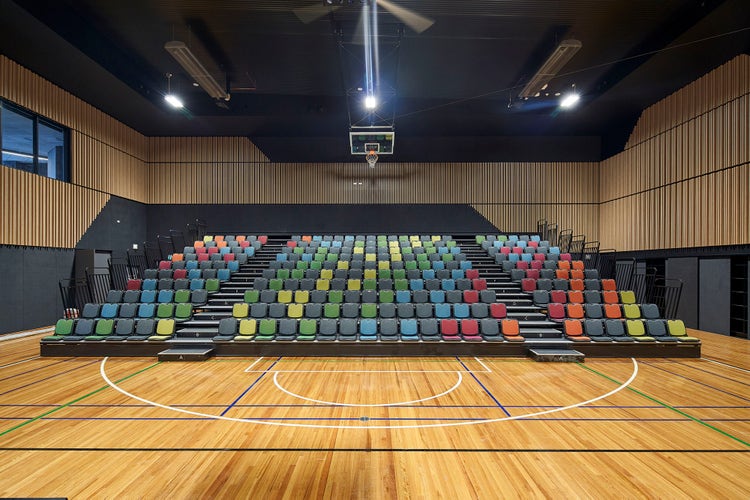 Multi-coloured tiered seating inside a large sports hall with wooden panelled walls and spotlights