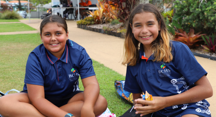 Two smiling students sitting on grass eating a snack