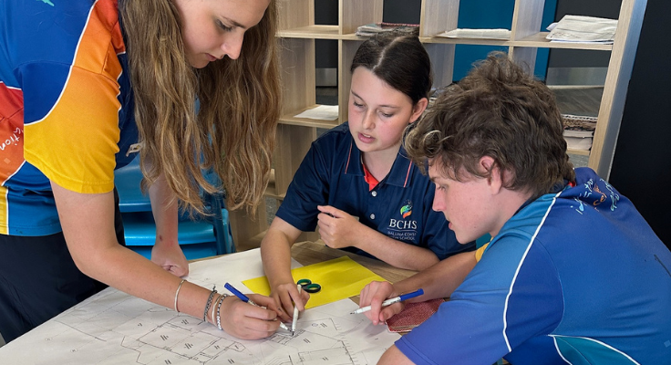 Three students around a table using pencils to point at architecture plans
