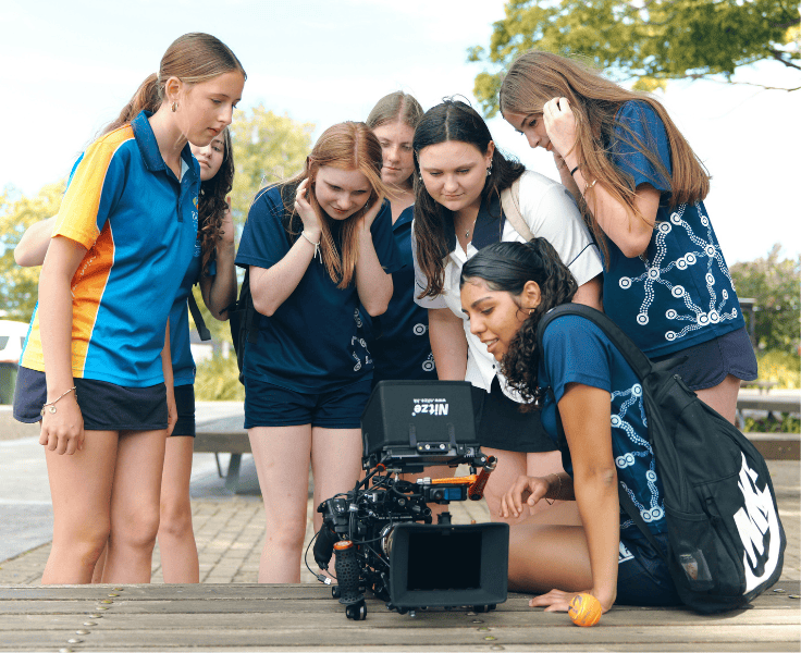 Six students gathered around a movie camera looking at it closely with interest.