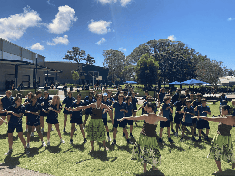 A large group of students in the playground having fun learning dance moves from the laughing female Polynesian dancers