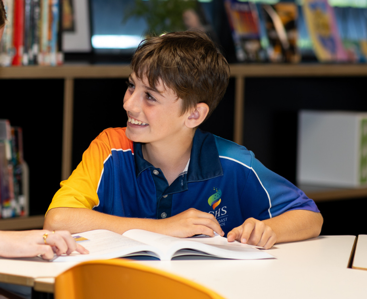 Junior student in library smiling at a person off-screen who is pointing at a page on his book