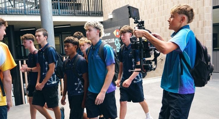 Male student using a large hand-held film camera to video a group of male students walking