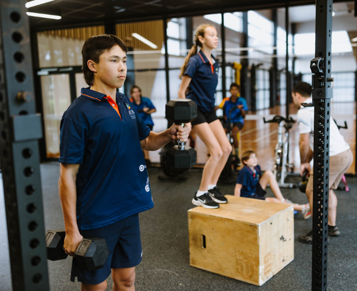 Students training inside the gym with male student using handweights in foreground, female student doing box jumps in mid shot and teacher instructing student on floor work in background