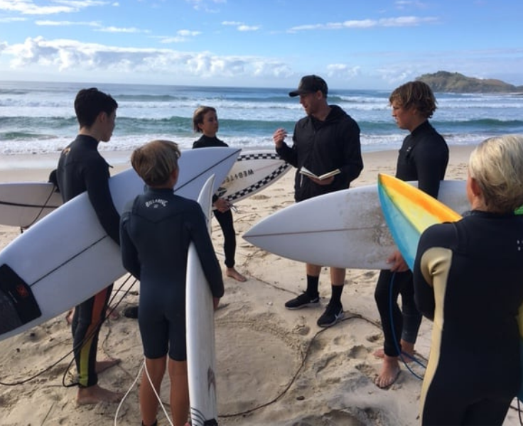 Group of surfers with boards under their hands gathered around an instructor at the beach