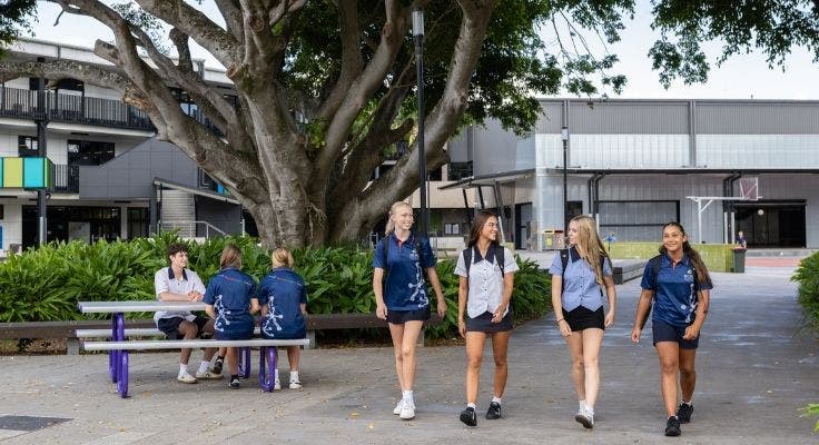 Smiling students walking in the playground while others area seated around a table under a large fig tree