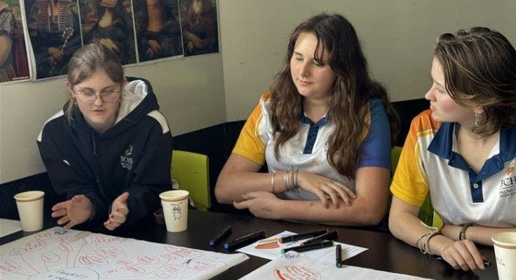 Three students engaged in discussion at a desk with brainstorming notes on butchers paper in front of them