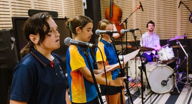 Music students standing in front of microphones singing in the foreground with music teacher on drums in background